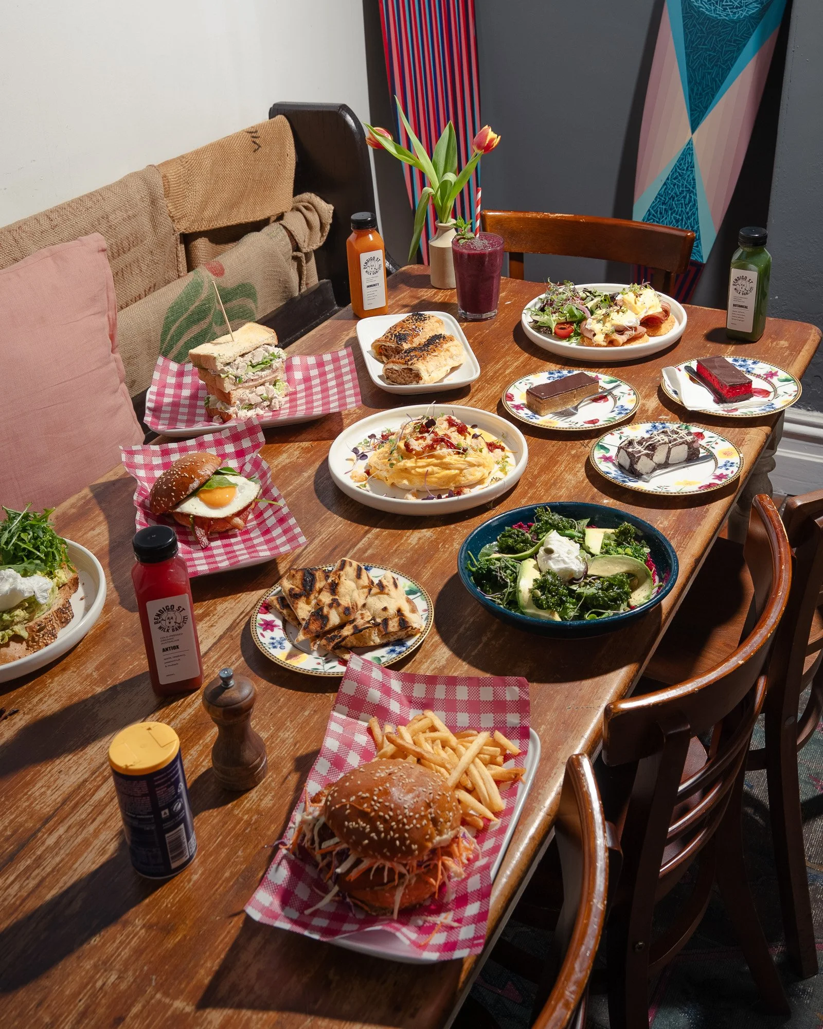 A wooden dining table filled with various plates of food, including sandwiches, fries, salads, pasta, and desserts, with bottles of sauces and drinks, in a cozy indoor setting with colorful decor and a flower vase.