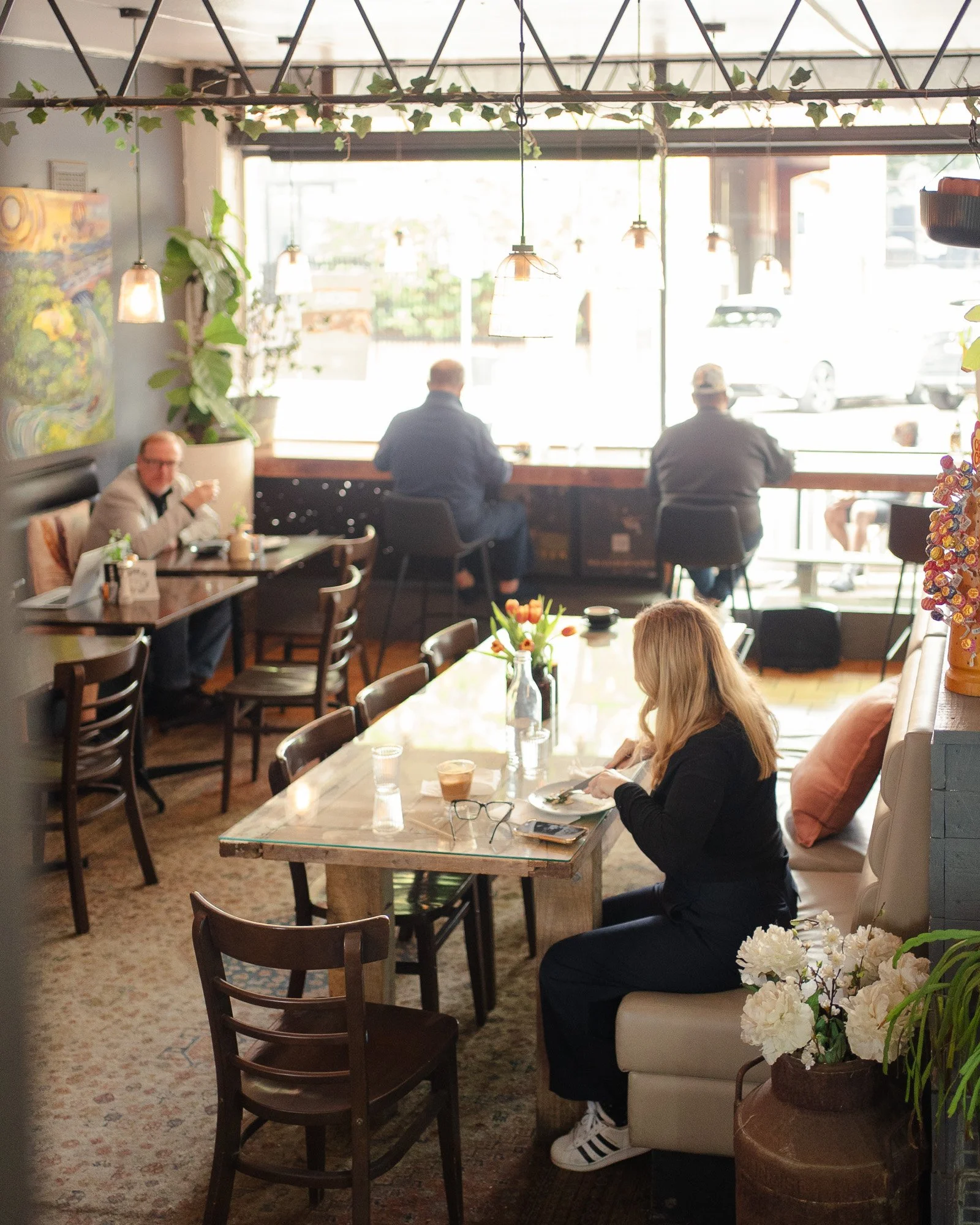 Interior of a cozy cafe with large windows, a few people sitting at tables and a booth, and decorative plants and artwork.
