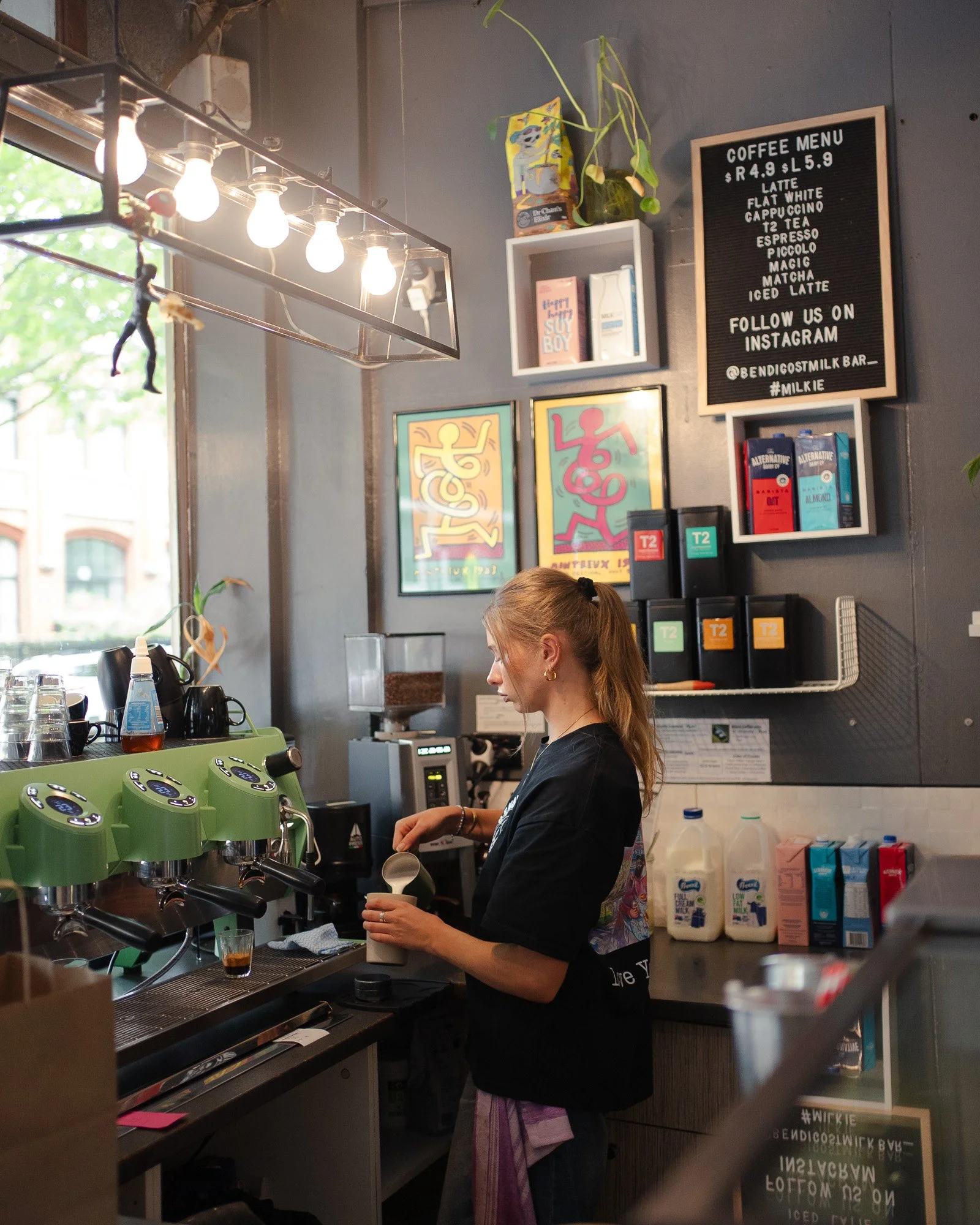 A barista prepares coffee behind a coffee bar in a modern cafe, with artwork on the wall and a black menu board displaying prices and drinks.