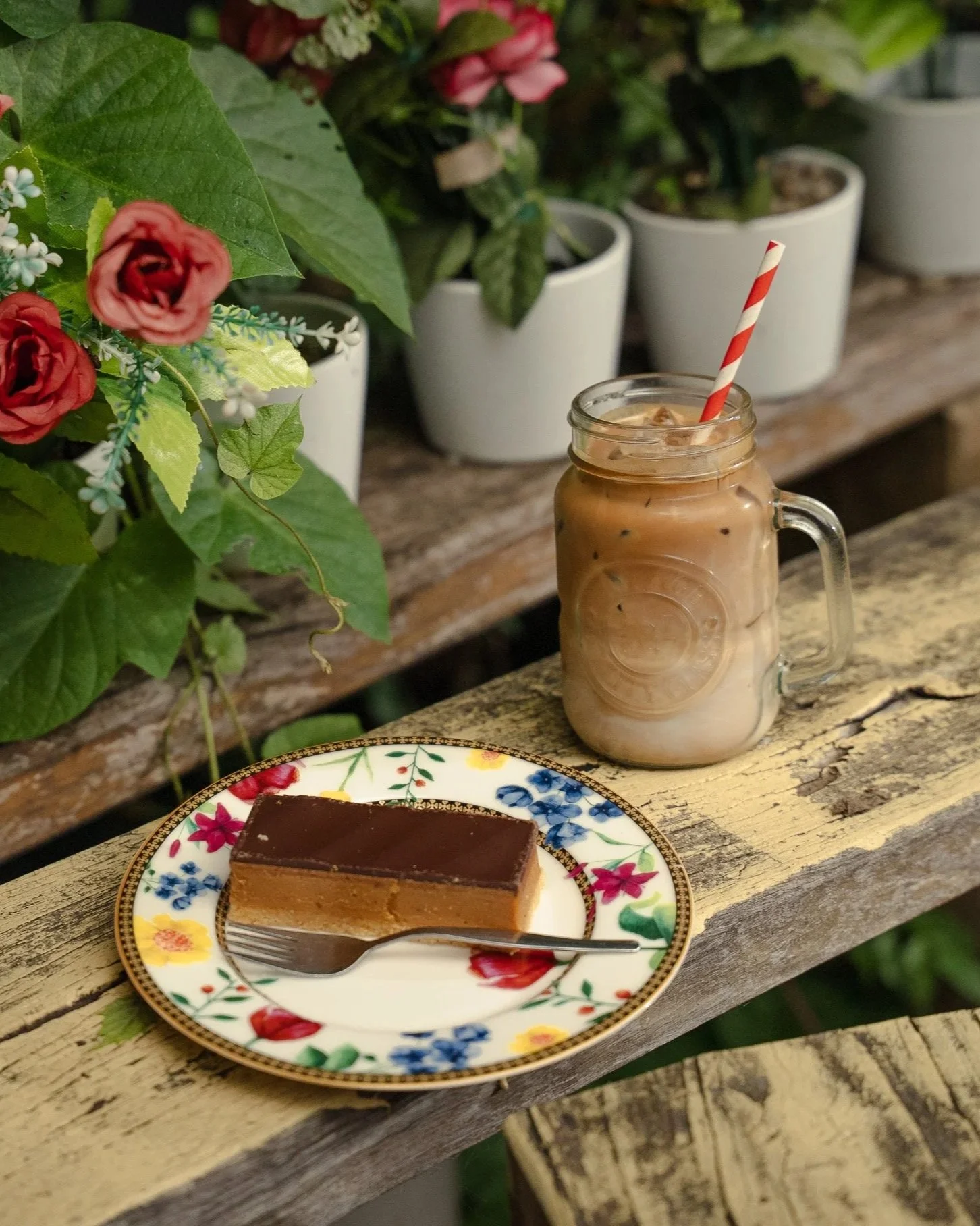 A plate with a piece of layered dessert, chocolate on top, on a rustic wooden table next to an iced coffee in a mason jar with a striped straw, with potted plants and pink flowers in the background.