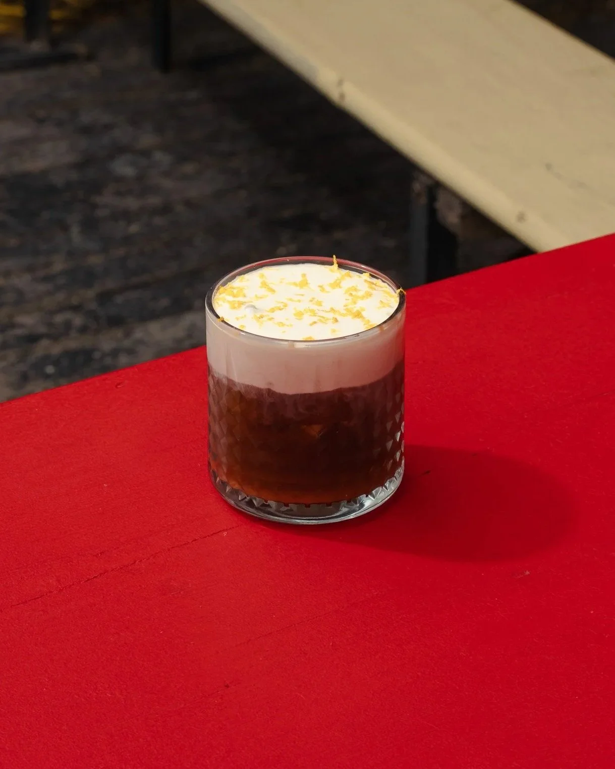 A glass of dark beer with a frothy head topped with lemon zest, placed on a red table.