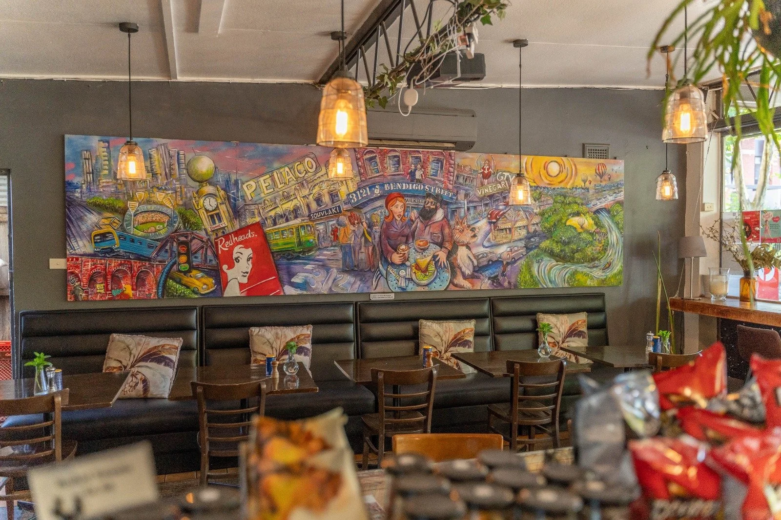Interior of a restaurant with a black booth, small tables, and decorative pillows. A colorful mural depicting city scenes, people, and landmarks hangs on the wall behind the booth. Hanging pendant lights illuminate the space, with plants and decorations adding to the ambiance.