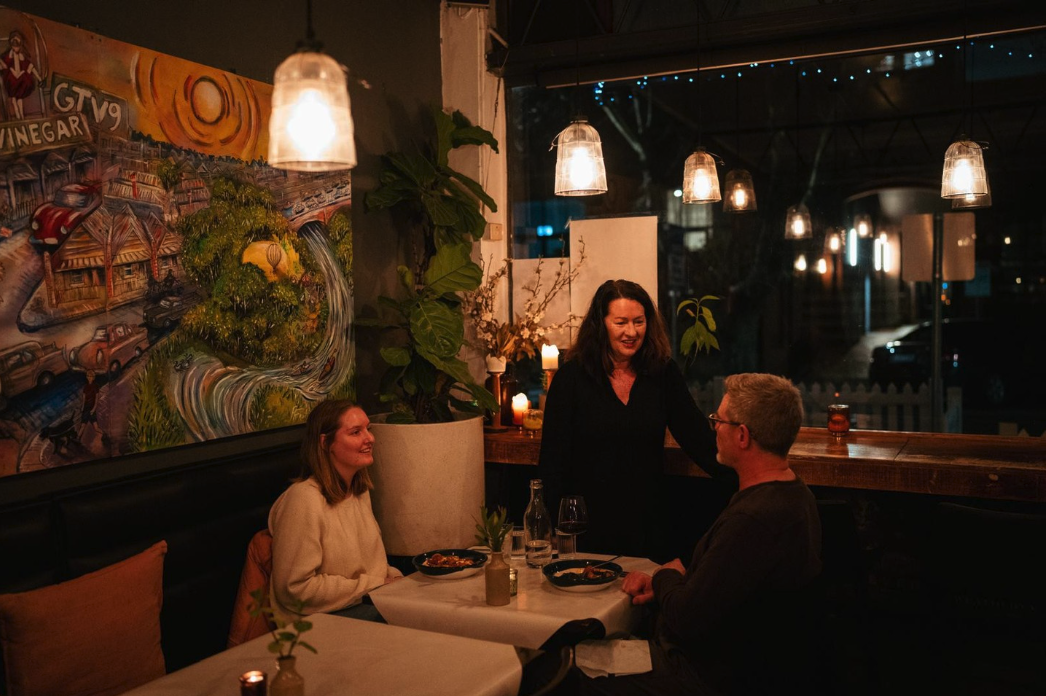 A woman with dark hair standing and talking to a couple at a restaurant table during the night. The woman is smiling, and there are candles, wine glasses, and plants on the table. The restaurant has a cozy, decorated interior with warm lighting and a large colorful mural on the wall.