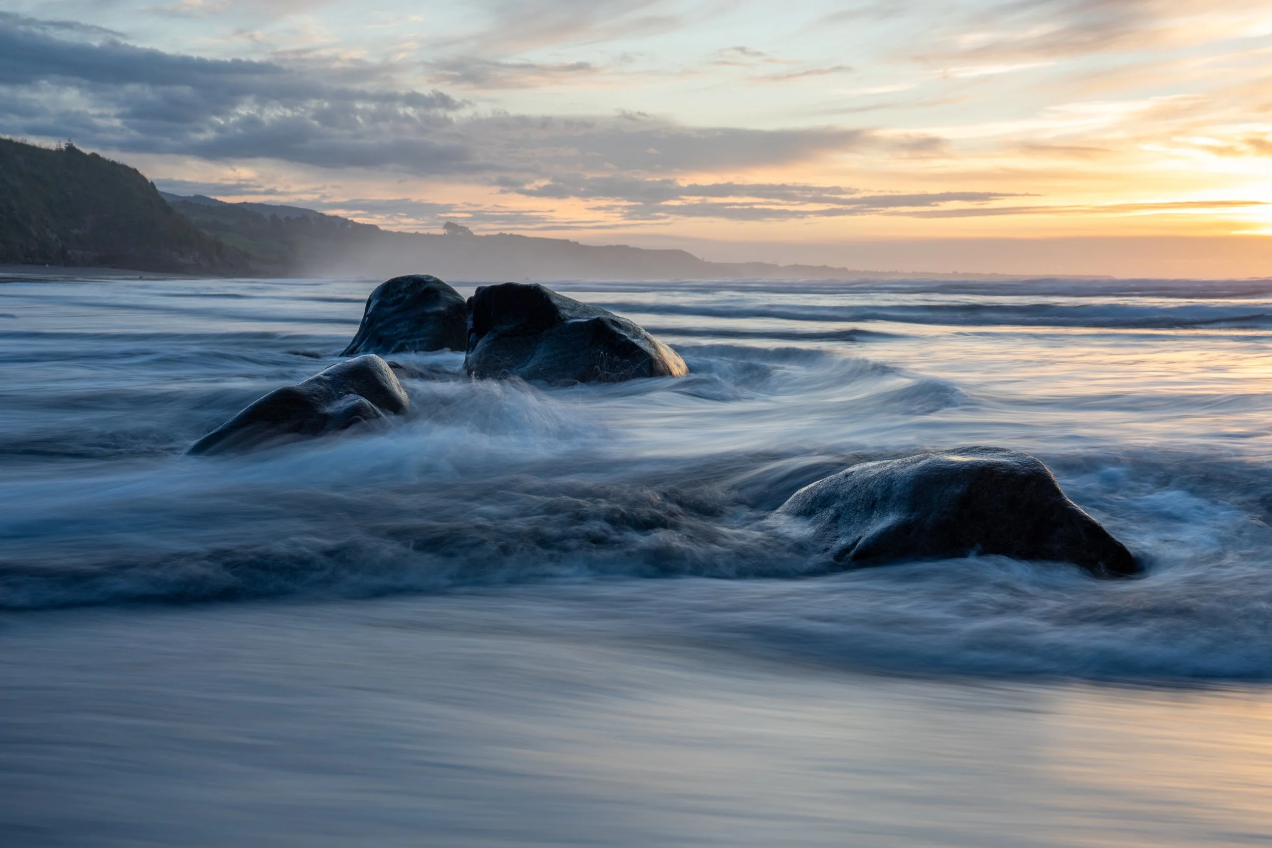 Sunset over ocean with rocks in the water and a cloudy sky.