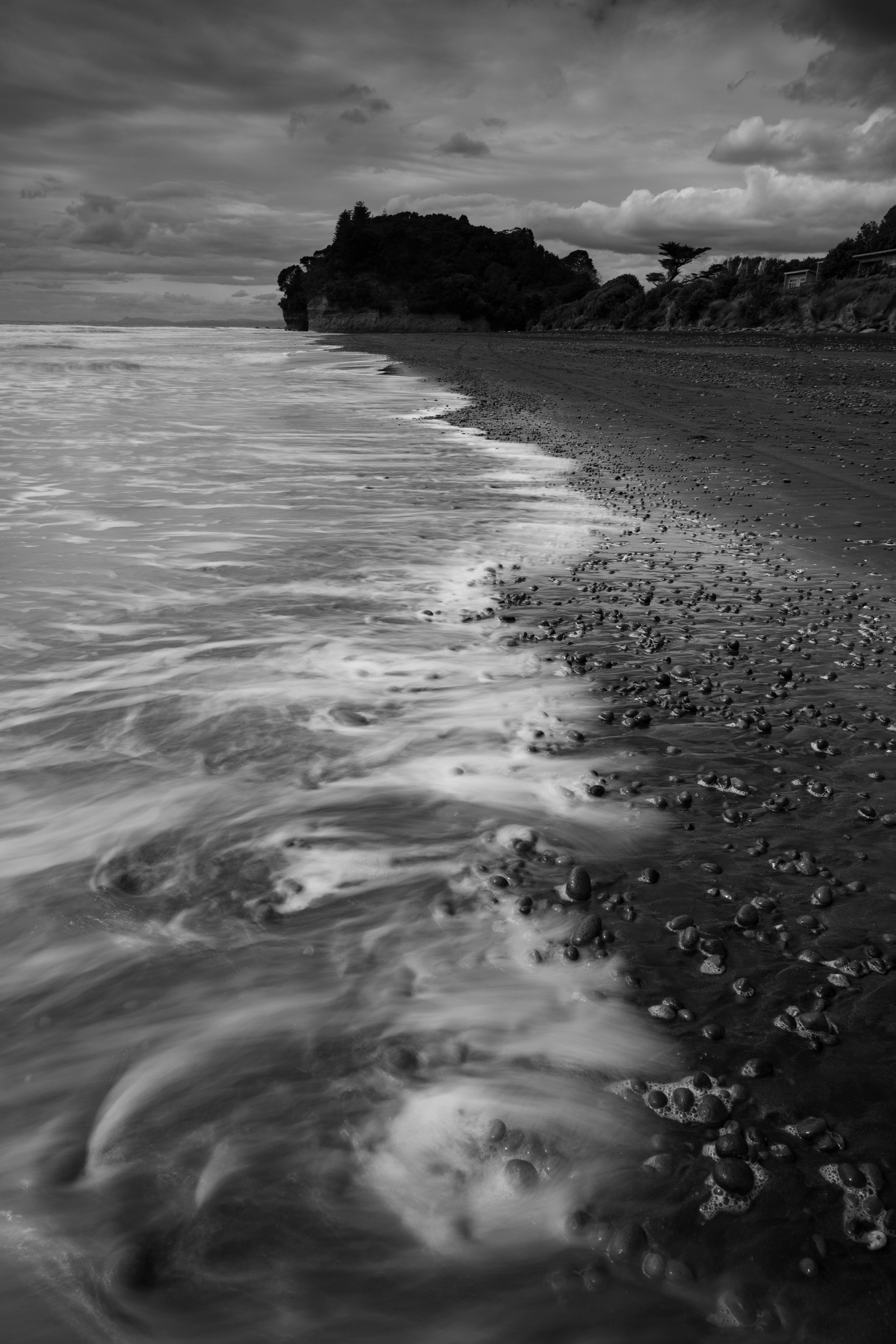 Black and white photo of a rocky beach with waves crashing and a wooded hillside in the background.