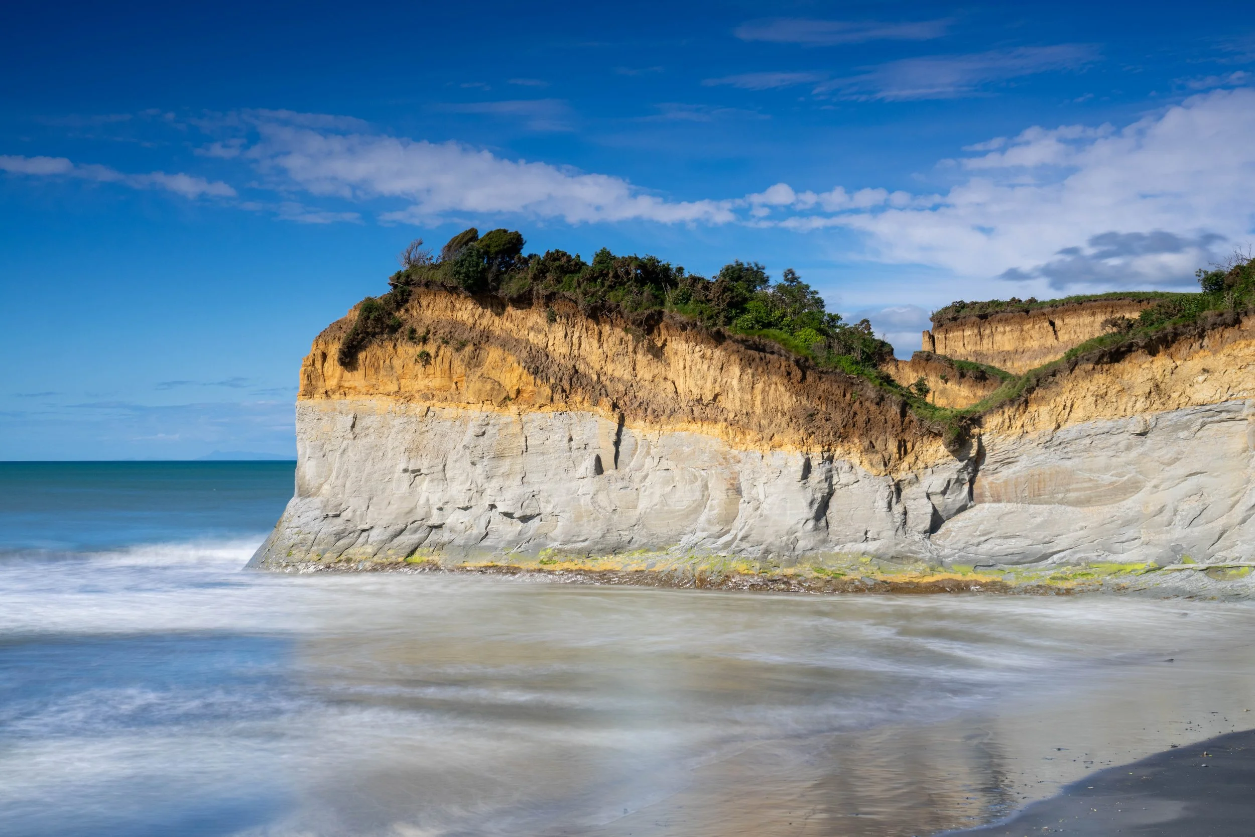Cliffside with layered rock formations along a beach, blue sky with clouds, and ocean waves in the foreground.