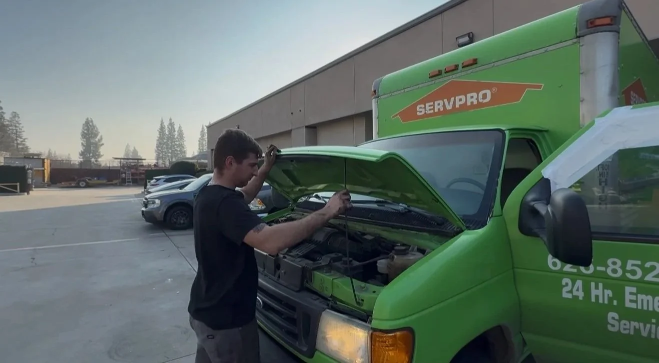 A man inspecting the engine of a green delivery van in a parking lot, with a row of cars and a warehouse in the background.