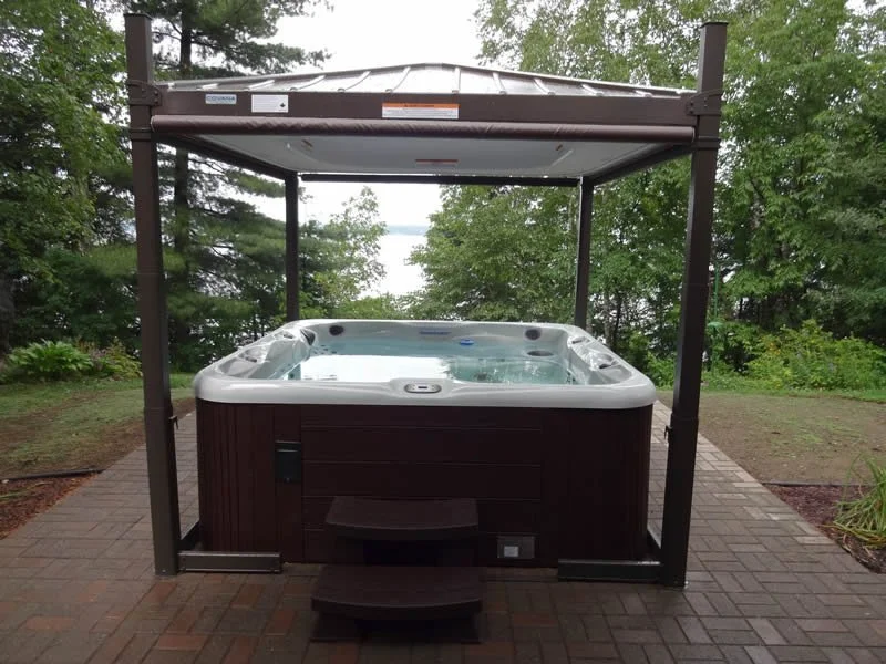Outdoor hot tub on a brick patio with a wooden canopy and lush green trees in the background.