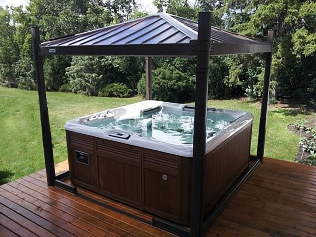 Hot tub in a backyard with a wooden deck and a black gazebo roof, surrounded by green trees and bushes.