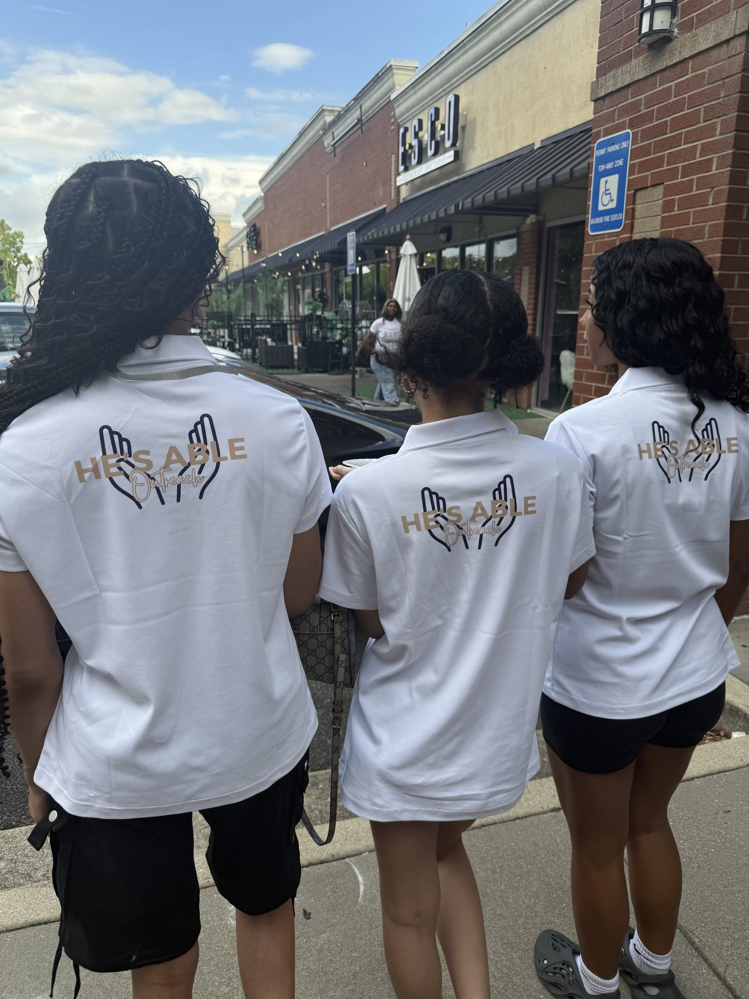 Three women wearing white shirts with logos that say "HE'S ABLE Outreach" on a sidewalk outside a restaurant named LSCO.