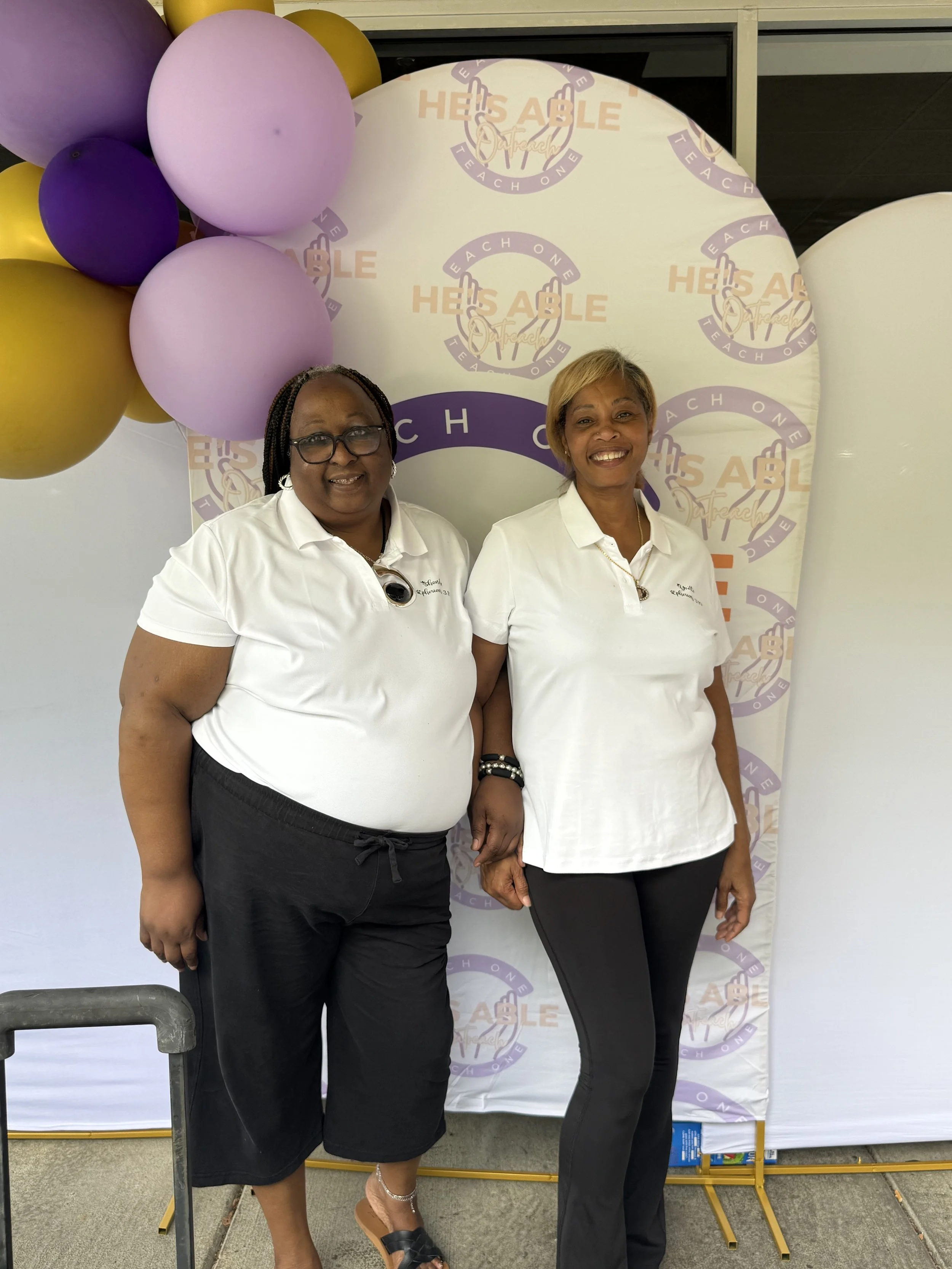 Two women standing side by side, smiling, with a backdrop and balloons. The backdrop has the text 'He's Able' and 'Teach One.' The women are wearing white shirts and black pants.