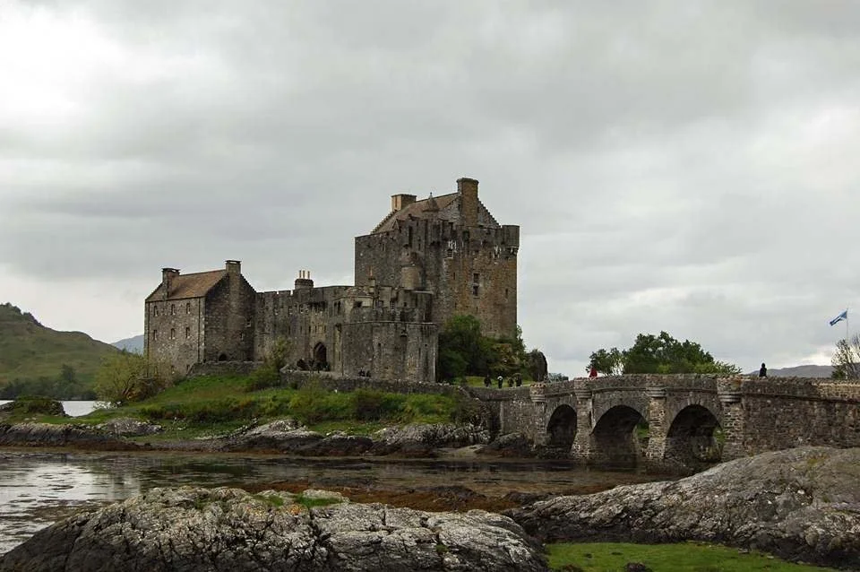 Eilean Donan Castle sits along the grass and a river with a grey sky in the background.
