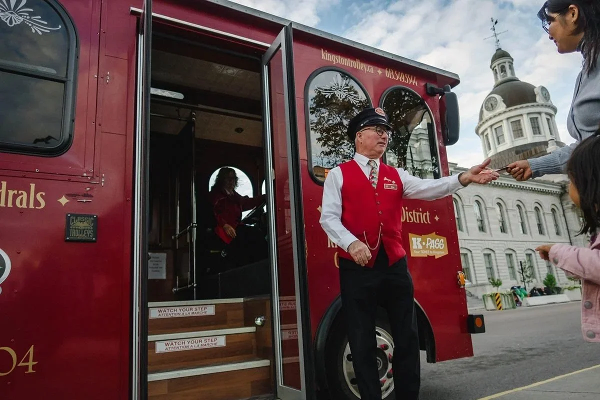 A trolley conductor dressed in a red and white suit hands a mom and daughter a ticket.