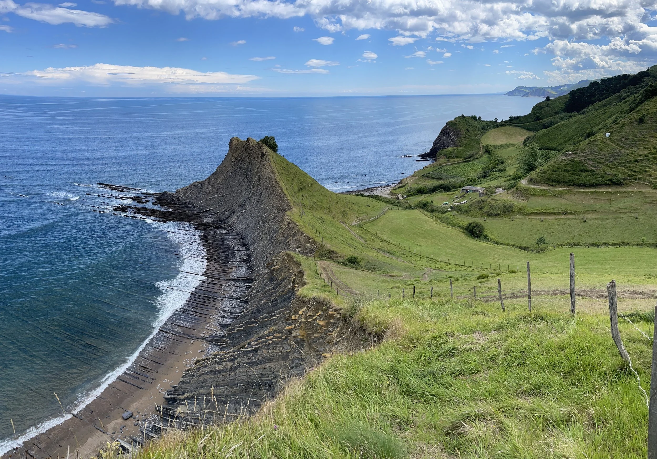 Coastal landscape with green grassy hills, rocky coastline, and a calm blue sea under a partly cloudy sky.