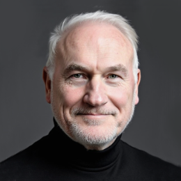 Portrait of a middle-aged man with white hair and beard, wearing a black turtleneck, smiling against a gray background.