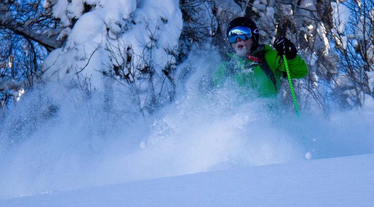 A heli skier cruises through powdered snow in a green ski jacket.