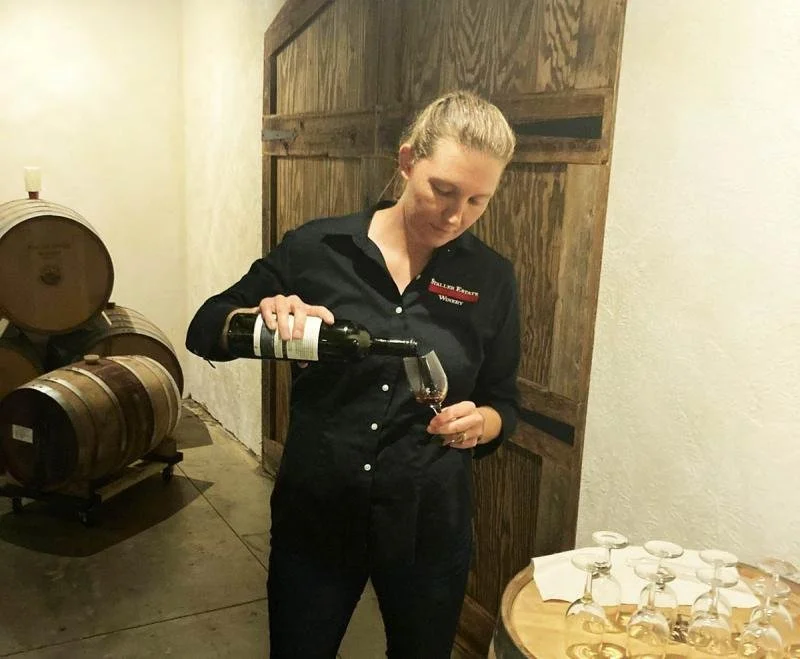 A woman in a black outfit pours wine into a bottle in a cellar.
