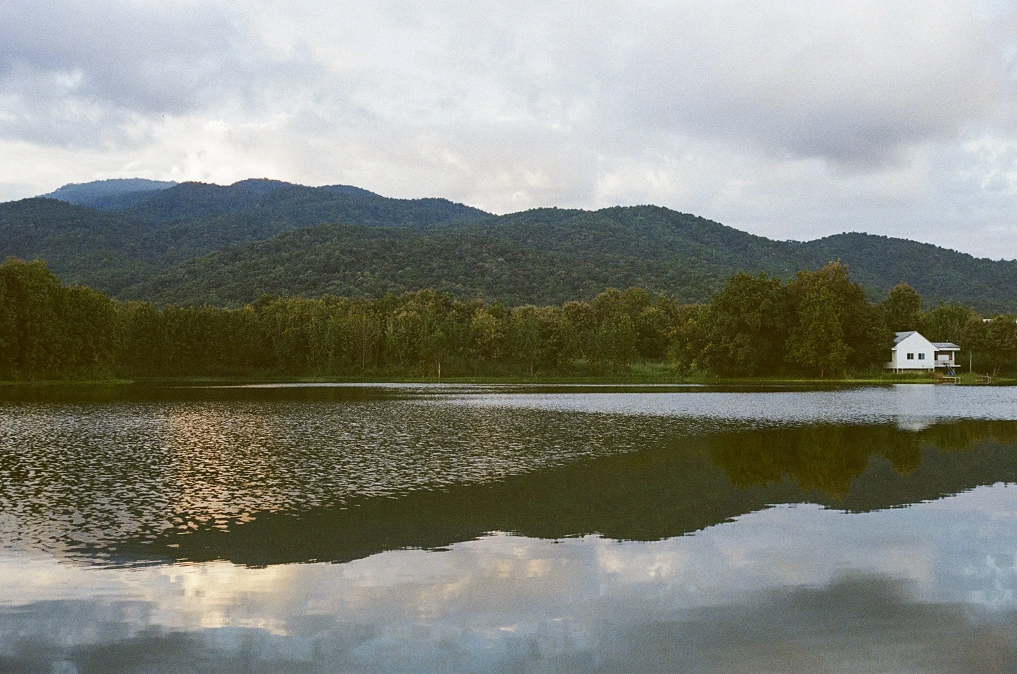 A house and Doi Suthep reflect in the water of Lake Aunjai during sunset.