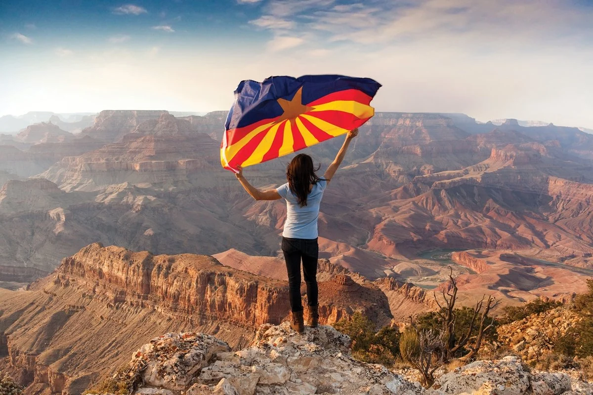 A woman in black jeans and a blue tea shirt holds an Arizona flag in the wind overlooking the Grand Canyon.