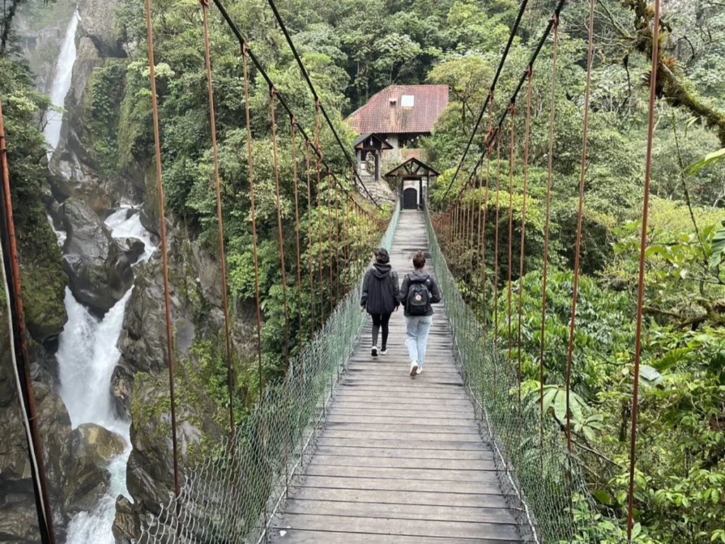 2 girls cross a bridge next to a rushing waterfall in Baños, Ecuador.