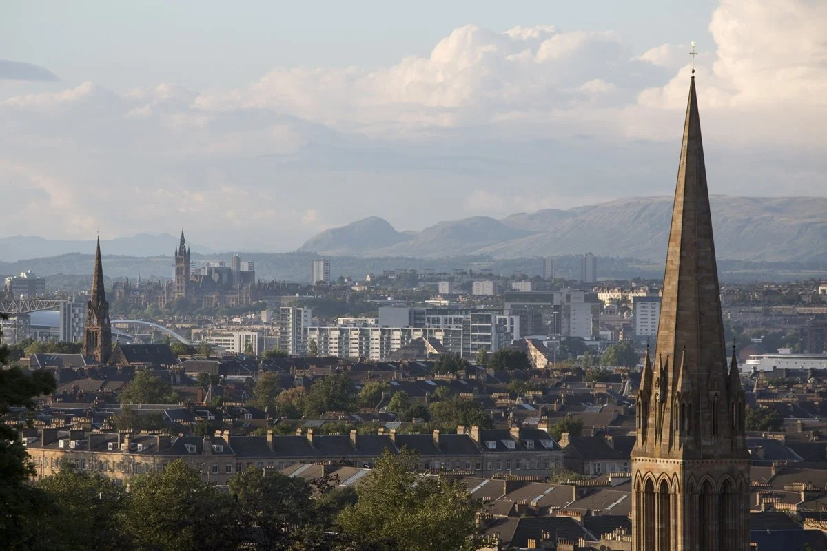 Panoramic view of Glasgow, Scotland, showing the city skyline with historic church spires, modern buildings, and the distant hills under a partly cloudy sky.