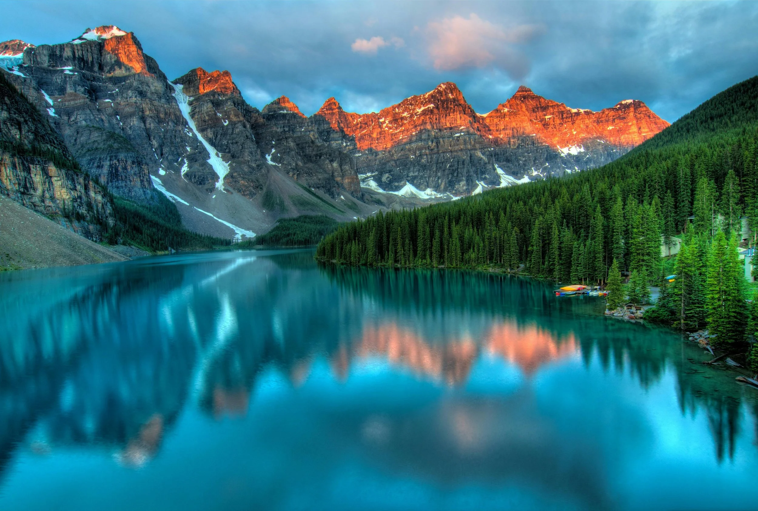 A scenic mountain landscape featuring a calm lake reflecting the snow-capped mountains, dense green pine forest, and colorful boats docked along the shoreline at sunset.