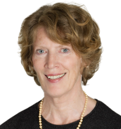 A smiling woman with short, curly, reddish-brown hair wearing a black top and pearl necklace