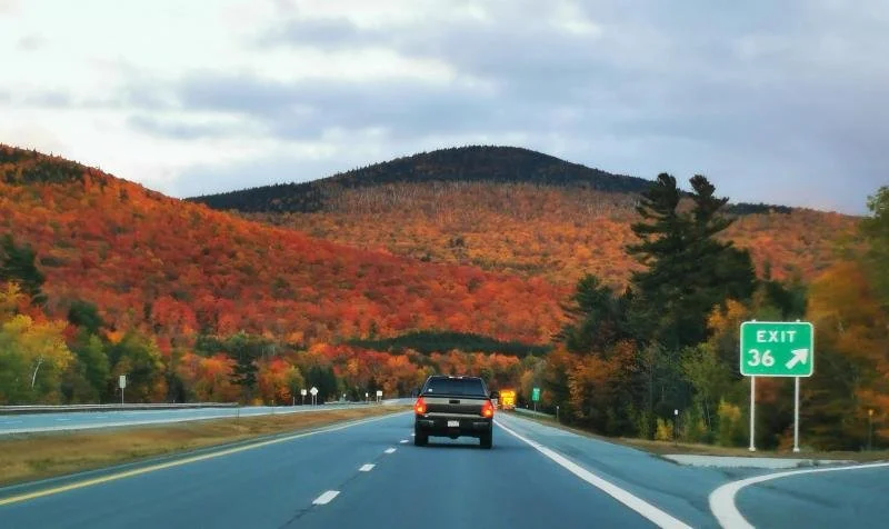 A black truck passes exit 36 in New Hampshire in front of a mountain full of bright fall trees.
