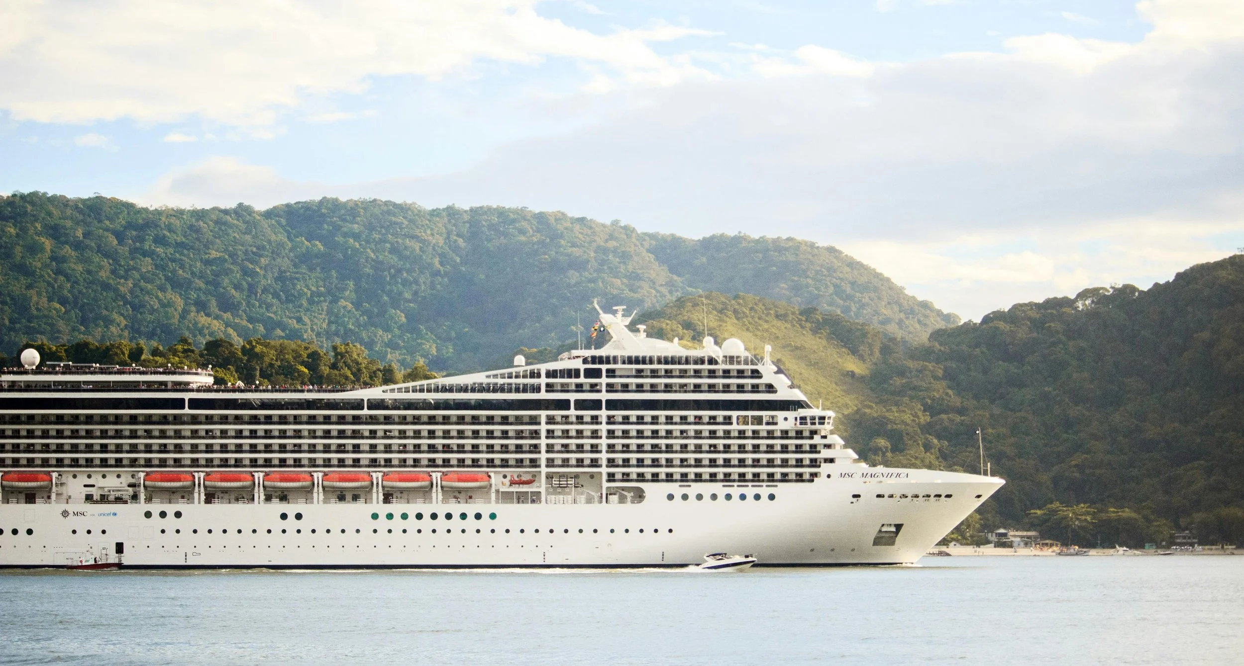 Large white cruise ship named MSC Magnifica sailing on water with green forested hills in the background.