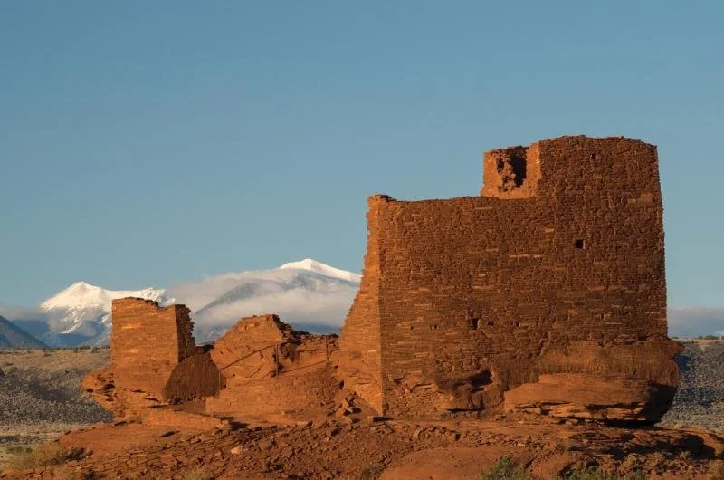 The red rock of the Wupatki National Monument stands in front of snow covered mountains.