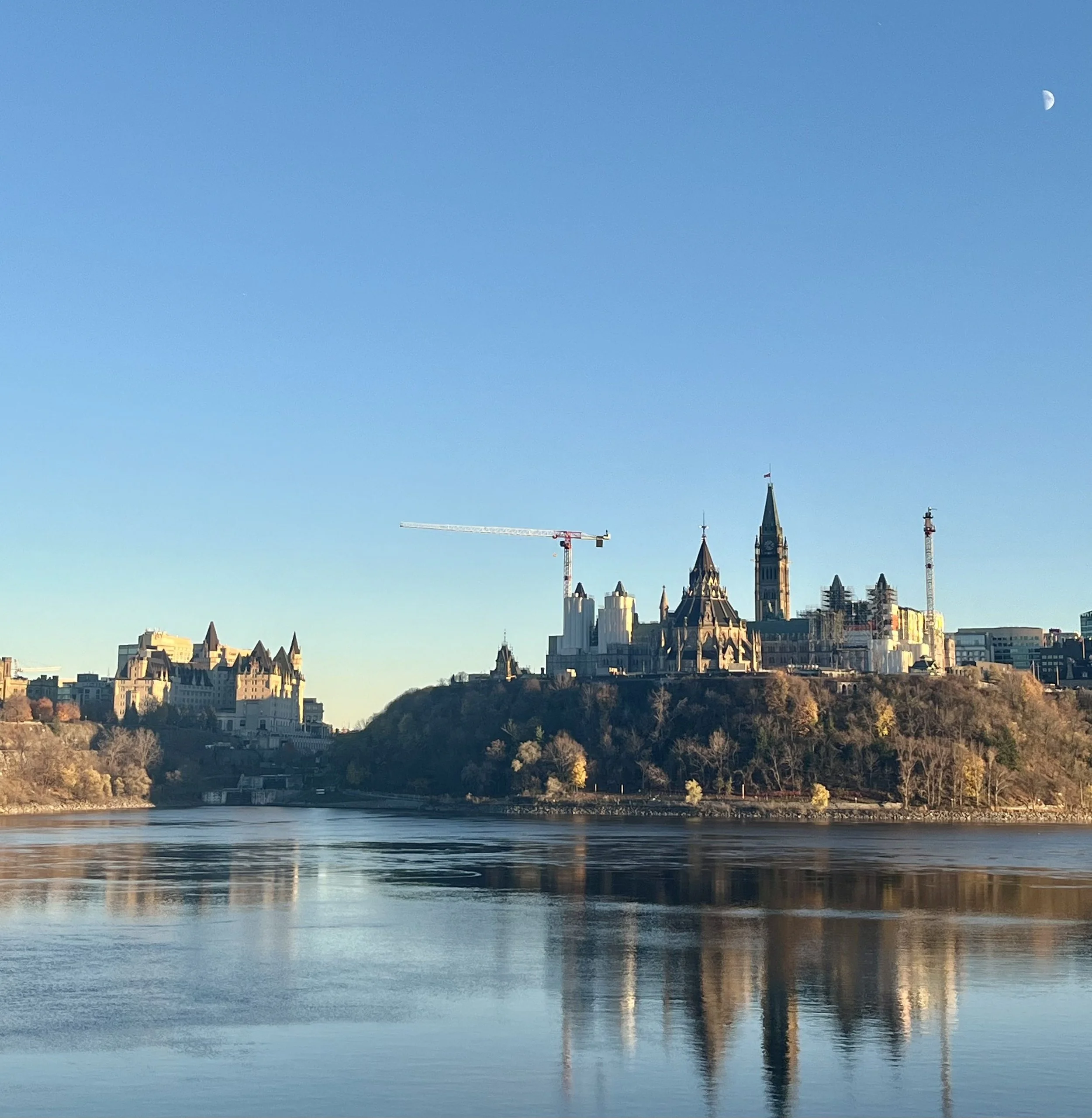 View of a city skyline with historic and modern buildings on a hill, a river in the foreground, and a clear blue sky with a moon visible.