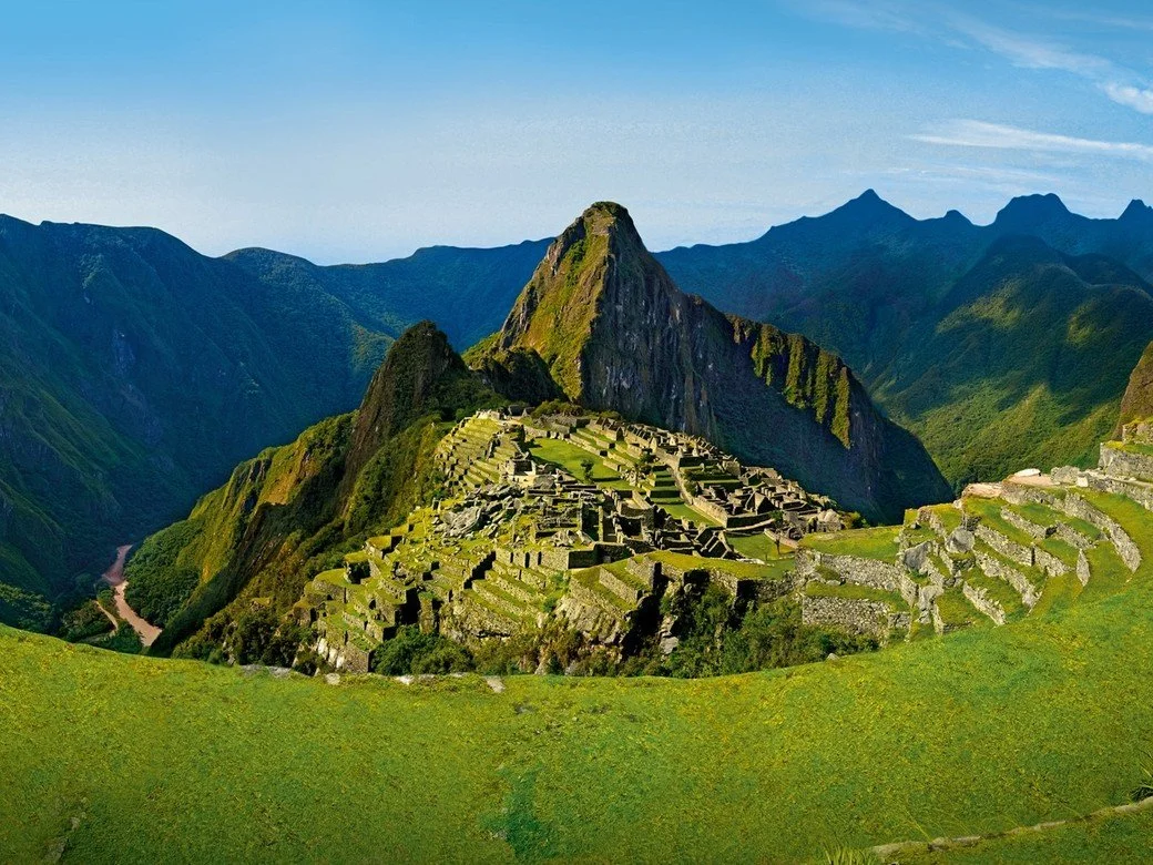 Machu Picchu surrounded by green fields, clear skies and mountains.