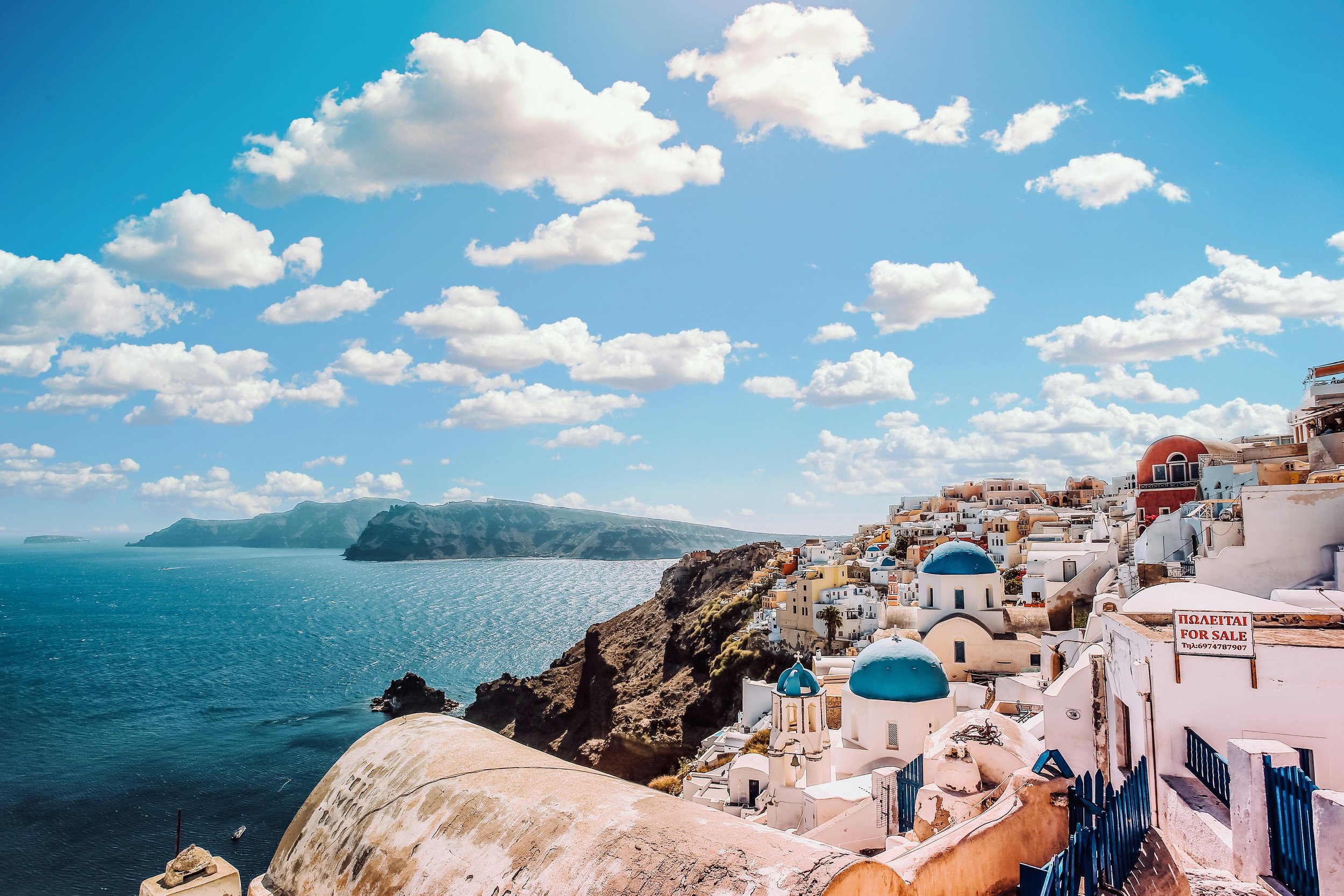 A coastal village with white buildings and blue domed roofs on a hillside overlooking the ocean, with a bright blue sky and scattered clouds.