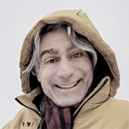 A smiling man with gray hair and a beard wearing a yellow jacket and a brown hood, looking into the camera.