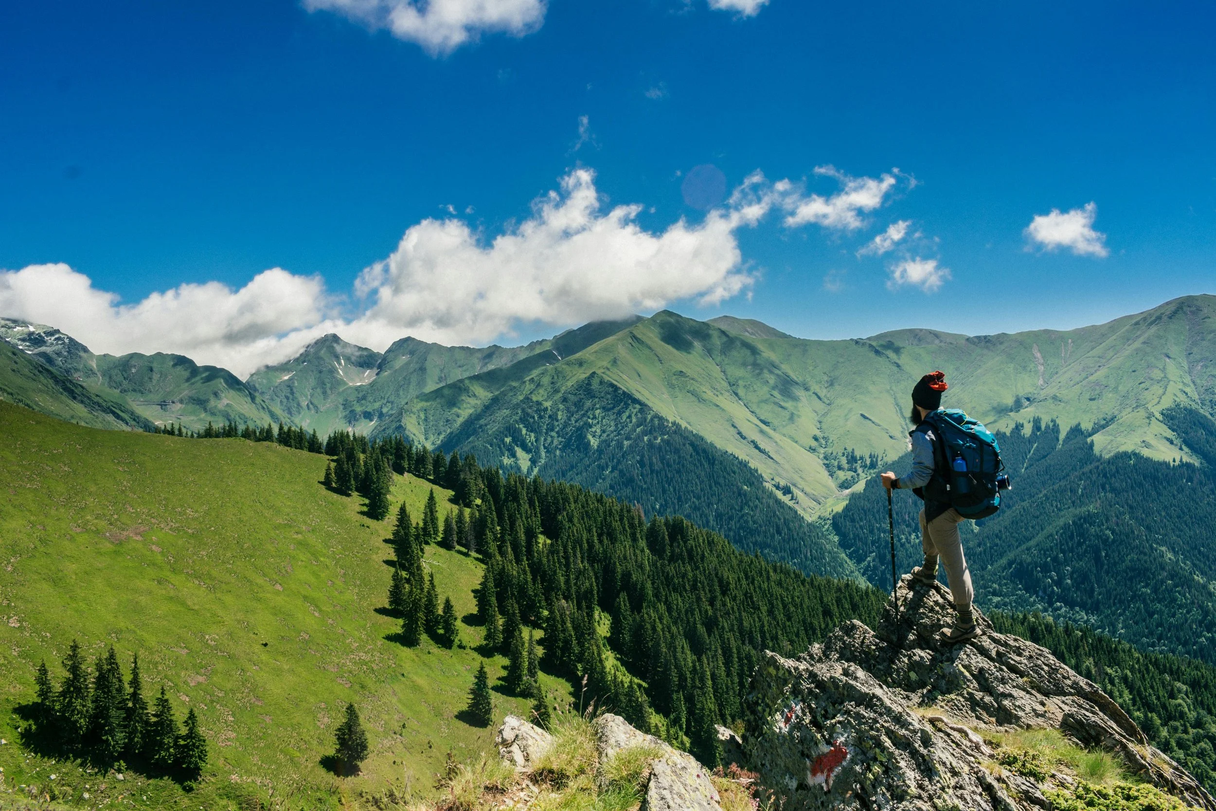 A hiker with a backpack and walking stick standing on a rocky outcrop, overlooking green mountainous landscape with trees and clouds in the sky.