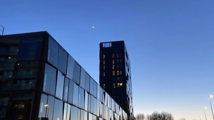 The moon towers over a glass building in Amsterdam during sunset.
