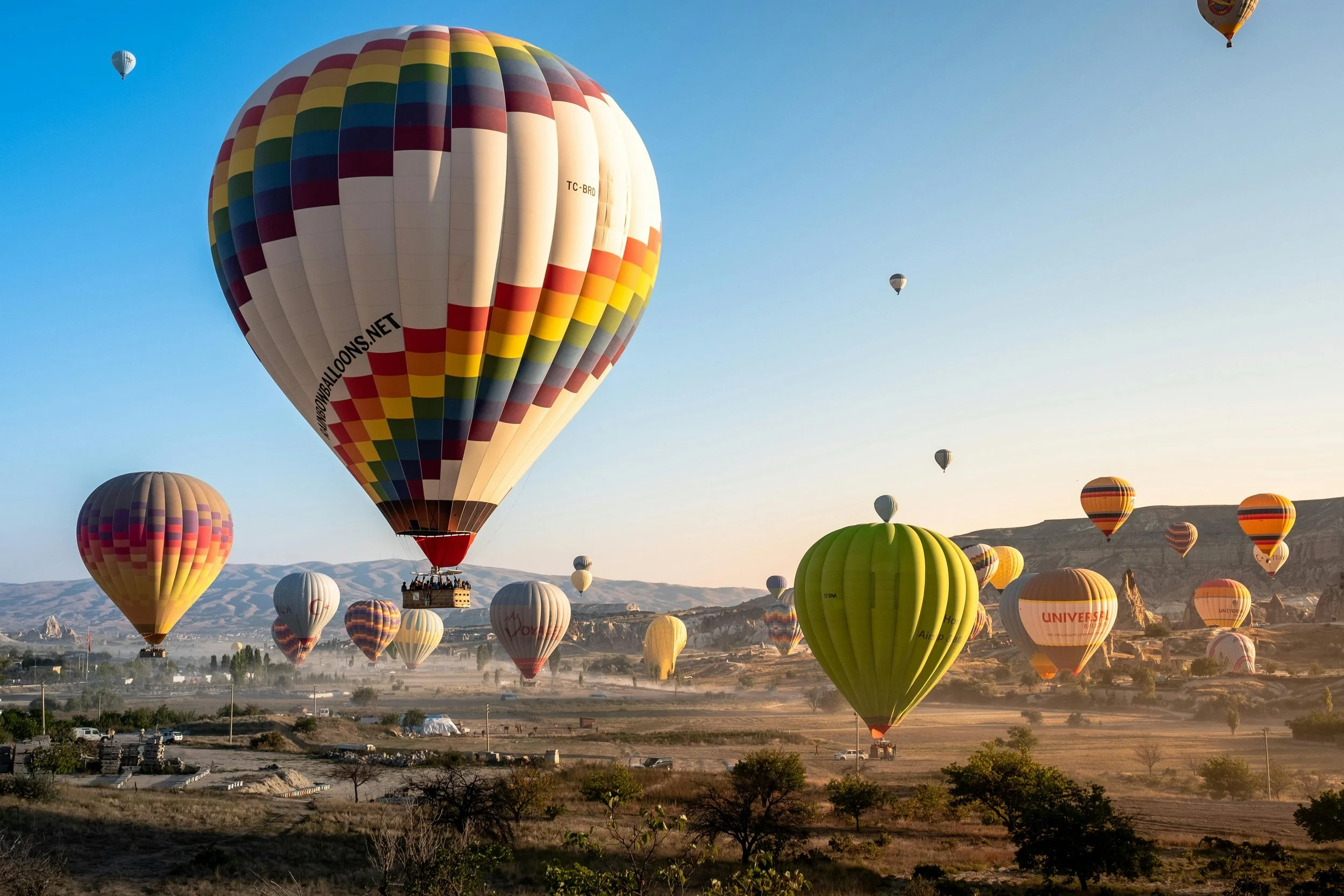 Multiple colorful hot air balloons floating in the sky over a rural landscape during sunrise or sunset.