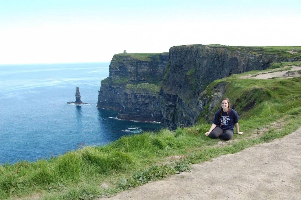A girl smiles on the cliffs of Moher where O'Brien's tower can be seen in the background.