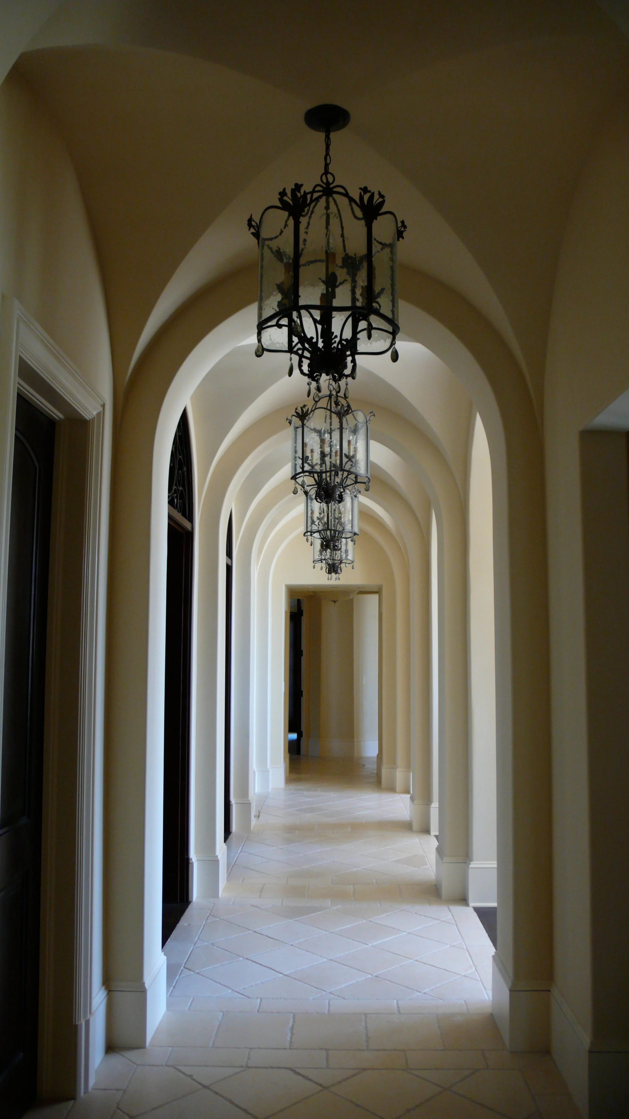 Elegant venetian plaster hallway with arched ceilings and hanging chandeliers, beige walls, white trim, tiled floor in a light color.