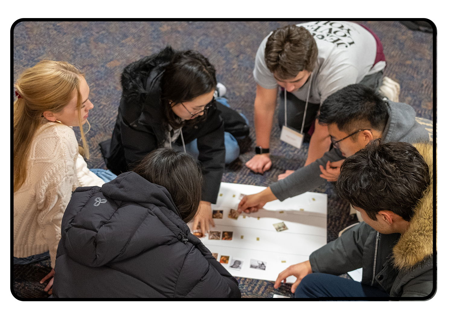 Group of six young people sitting and kneeling on a carpeted floor, working together on a large sheet with small photographs and sticky notes, engaged in a collaborative activity.