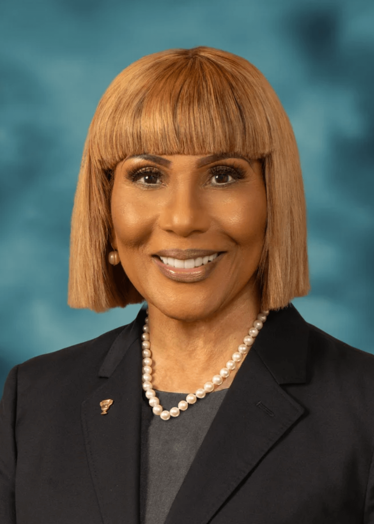 A professional woman with a shoulder-length ginger bob haircut, wearing a dark blazer, pearl necklace, and earrings, smiling against a blue background.