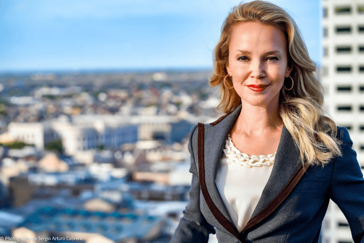A woman with long blonde hair wearing a gray blazer over a white blouse, standing outdoors in front of a cityscape with buildings under a clear blue sky.