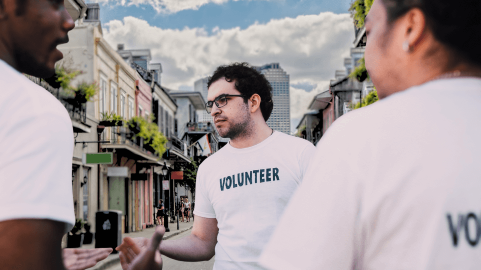 A man wearing glasses and a white volunteer shirt talking to two people on a city street during the day.