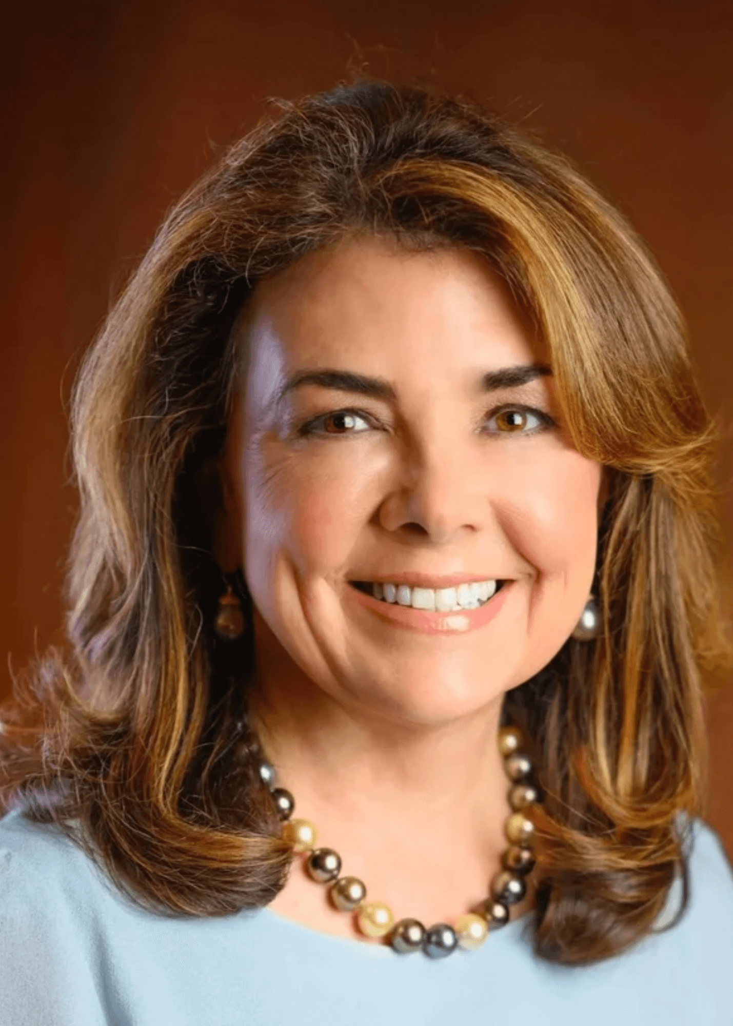 A woman with brown hair, wearing a light blue top, pearl necklace, and pearl earrings, smiling against a warm background.