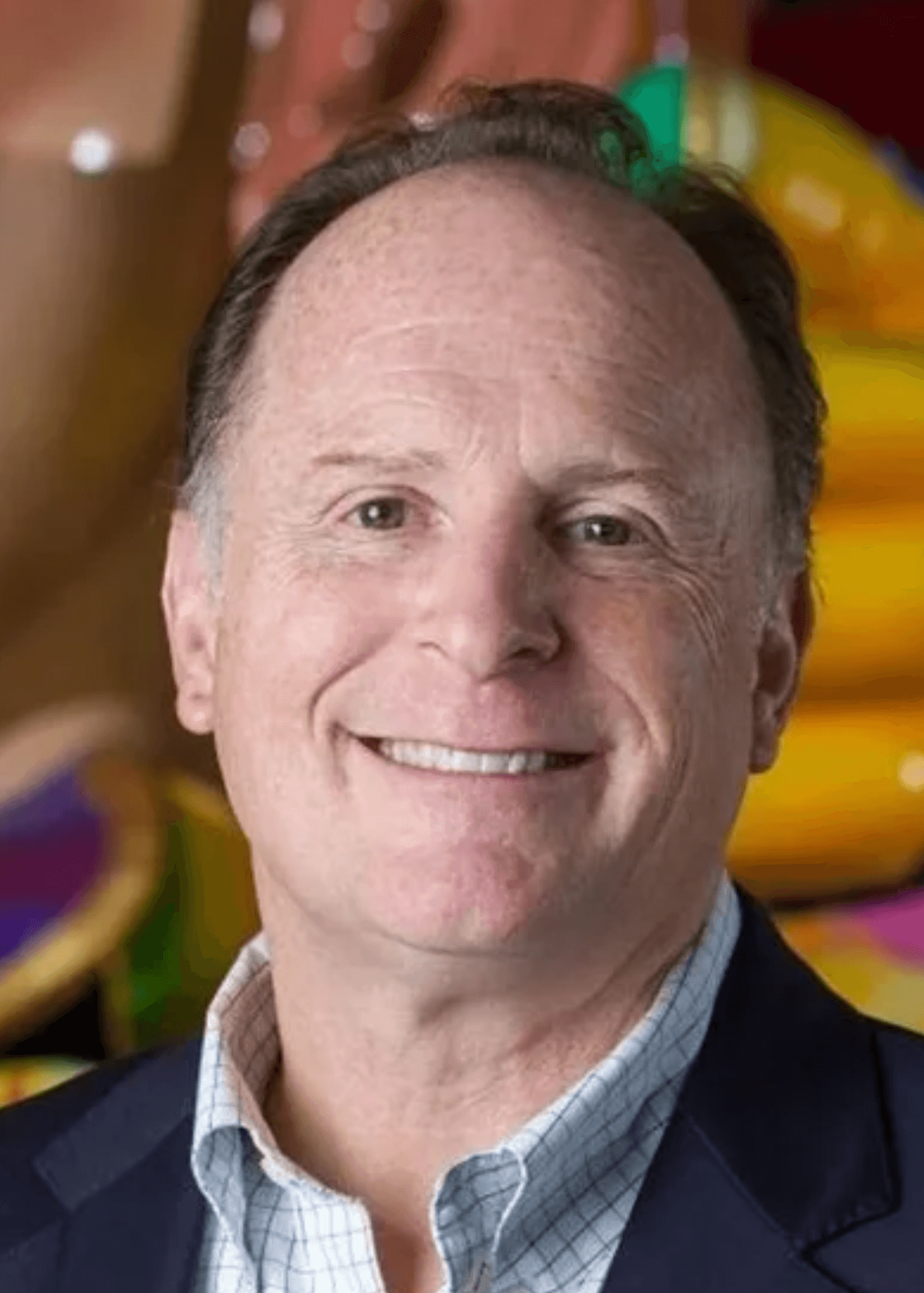Close-up portrait of smiling middle-aged man with dark hair, wearing a checkered shirt and dark blazer, in front of colorful blurred background.