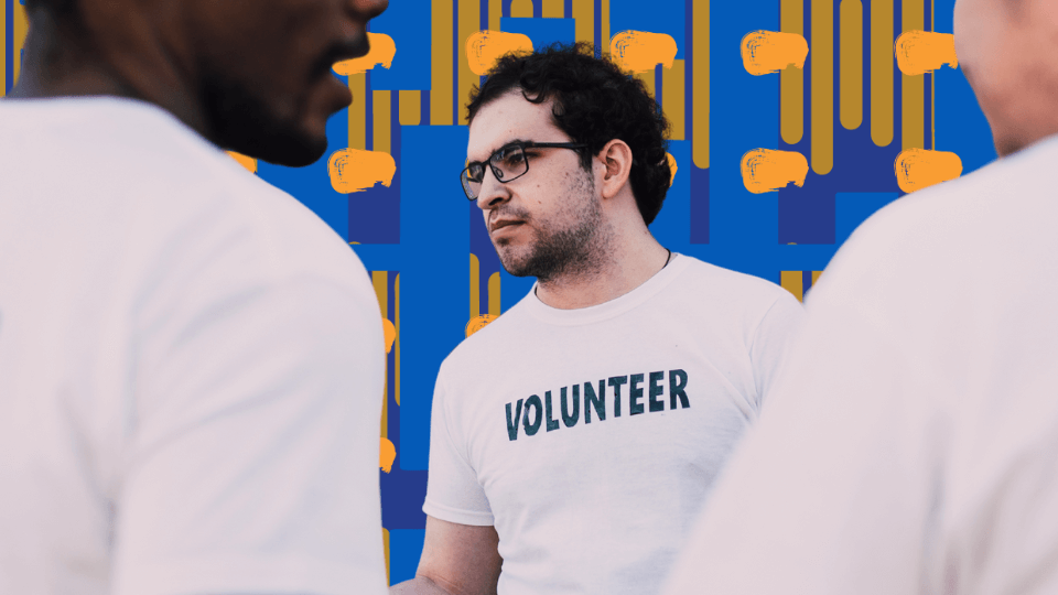Three people, two with their backs to the camera, talking to a man wearing glasses and a white shirt with 'VOLUNTEER' printed on it, on a city street with colorful buildings, shops, and a cloudy sky in the background.