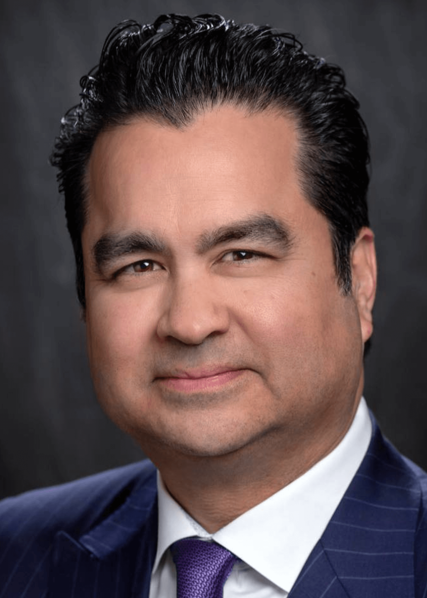 Close-up of a man with dark, slicked-back hair wearing a pinstripe suit, white shirt, and purple tie, looking at the camera with a neutral expression.