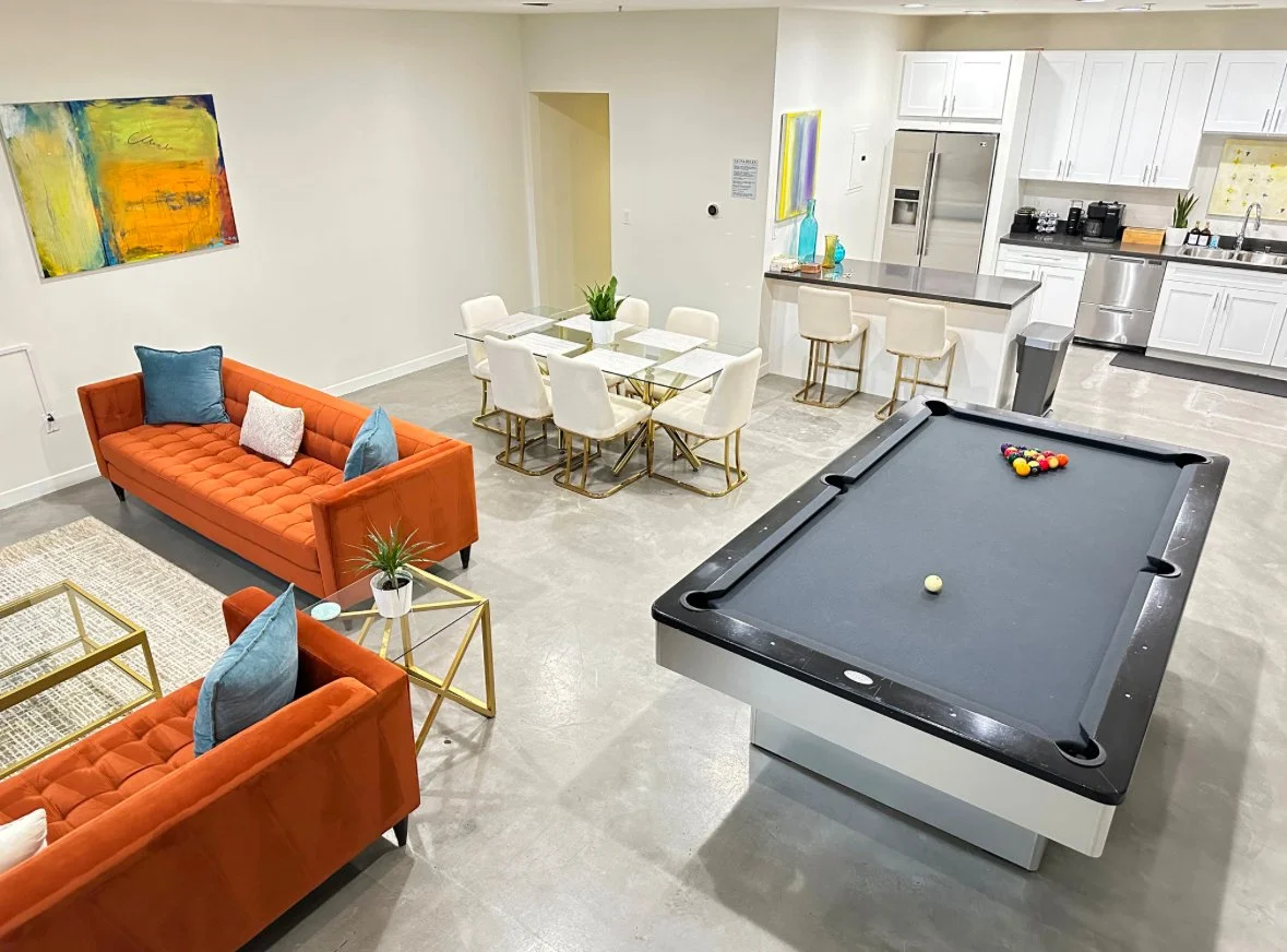Living room and kitchen area with orange sofas, blue pillows, a pool table, a dining table with white chairs, and white kitchen cabinets with stainless steel appliances.