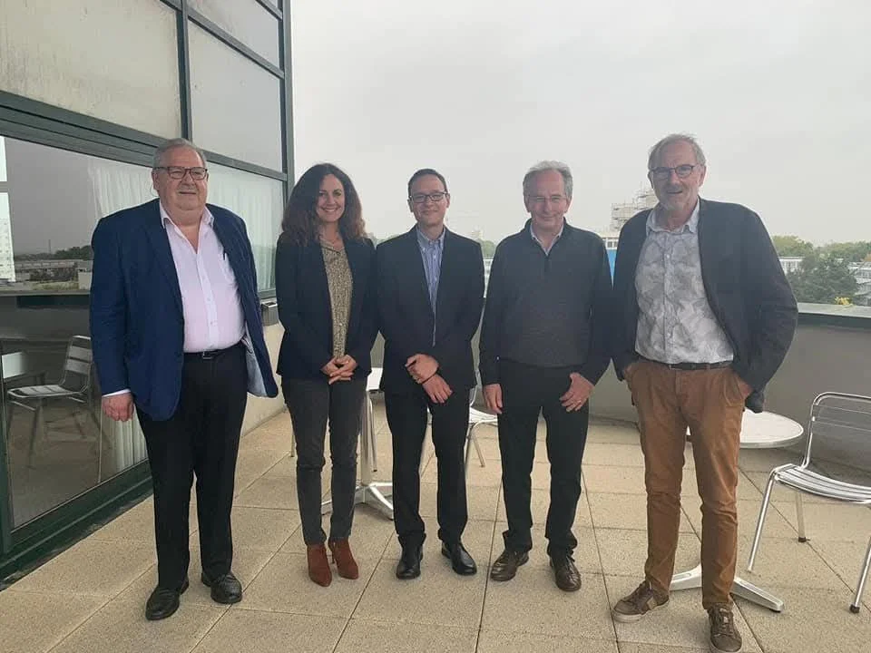 Cinco personas en ropa de negocios de pie en una terraza con vista a la ciudad, sonriendo para la foto.