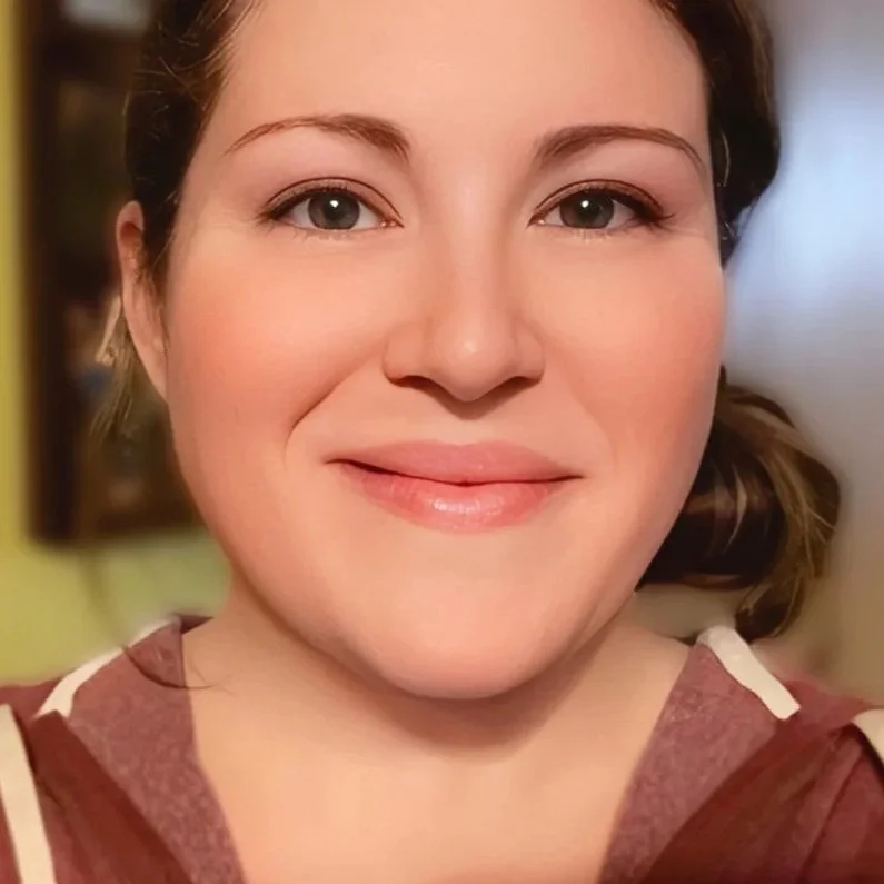 Close-up of a smiling woman with brown hair, wearing a maroon hoodie, indoors with a yellow wall in the background.